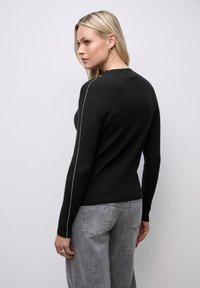 Czarny sweter dziany o ryflowanej fakturze, z białymi szwami bocznymi. Noszony z szarymi spodniami typu wide-leg, pokazany z tyłu.