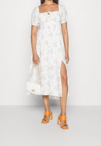 Robe blanche à fleurs avec des manches courtes bouffantes, un détail en forme de trou de serrure et une fente sur le côté. Associée à des sandales à talons blocs orange et un sac à main blanc structuré.