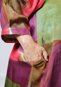 Erwachsene Hand, die in der Tasche eines bunten, gemusterten Kleidungsstücks mit Schattierungen von Rot, Lila, Grün und Braun ruht.