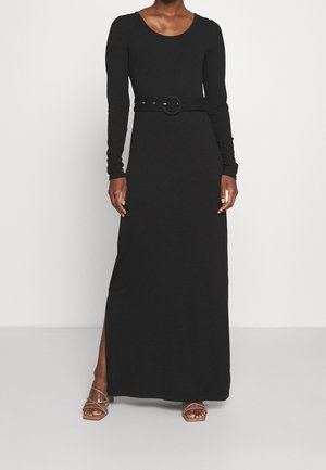 Kobieta ubrana w czarną długą sukienkę z długim rękawem, z okrągłym dekoltem i czarnym paskiem z okrągłą klamrą, w połączeniu z brązowymi sandałami na paski.