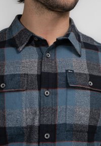 Camisa de franela de cuadros en tonos azules, grises y negros con detalles rojos. Con cuello abotonado y dos bolsillos frontales.