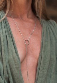 Collier en chaîne argentée avec un pendentif circulaire suspendu sur la poitrine, porté sur un vêtement vert texturé à col en V plongeant.