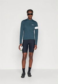 Sykkelejakke i mørk teal, hel glidelås, lange ermer med hvit aksent. Svarte sykkelshorts. Holder en grønn vannflaske. Sporty design.