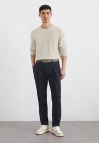 Beige textuur langemouwen overhemd, marineblauwe broek, bruine riem en beige sneakers met lichte accenten, tegen een eenvoudige witte achtergrond.