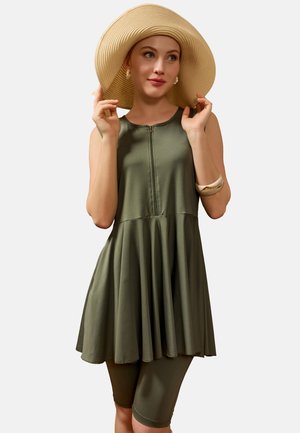 Donna che indossa un vestito verde oliva senza maniche con cerniera frontale e pantaloncini da bici abbinati, grande cappello da sole beige, orecchini di perle e braccialetto bianco.