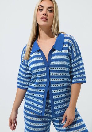 Blondine draagt een bijpassende blauw gestreepte gebreide cardigan en shorts met oogjesdetails tegen een effen lichte achtergrond.