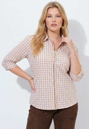 Mujer con el pelo largo y rubio que lleva una camisa de cuadros beige y blanca y pantalones marrones, posando con una mano en la cadera y la otra cerca del cuello.