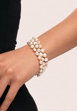Bracelet en perles avec un design superposé mettant en valeur des perles blanches de formes irrégulières et des accents dorés, porté sur un poignet contre une tenue sombre.