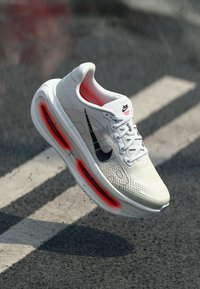 Λευκό αθλητικό παπούτσι με υφή στο πάνω μέρος, μαύρο λογότυπο της Nike και κόκκινη αερομονωτική σόλα. Είναι ορατά τα κορδόνια και η επενδεδυμένη κολάρο.