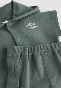 Grüne Baumwollkapuzenjacke mit Knopfleiste und gesticktem "kleiner Mann"-Text, kombiniert mit passenden grünen Baumwollhosen mit elastischem Bund.