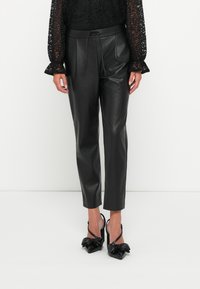 Zwarte faux leren broek met een taps toelopende pasvorm, voorzien van een trekkoord in de taille en een gladde textuur. Gekleed met zwarte pumps met strikaccenten.
