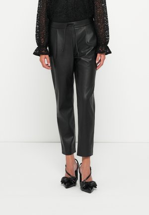 Zwarte faux leren broek met een taps toelopende pasvorm, voorzien van een trekkoord in de taille en een gladde textuur. Gekleed met zwarte pumps met strikaccenten.