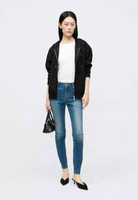 Sweatshirt com capuz preto, camisa justa branca, jeans skinny azuis, sapatos de salto alto pretos e uma pequena bolsa preta.