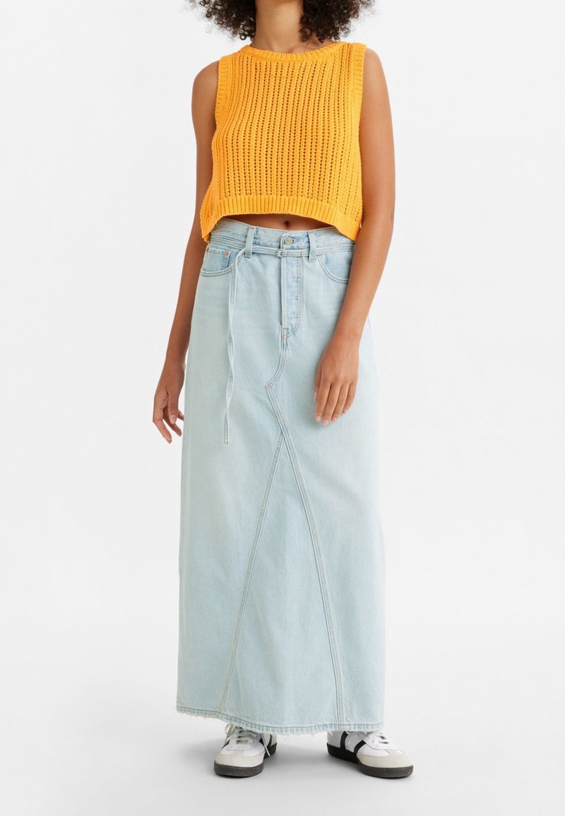 Femme portant un crop top orange sans manches en maille, une longue jupe en jean taille haute de couleur bleu clair et des baskets noires et blanches.