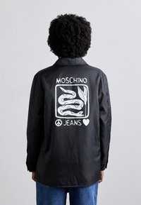 Zwarte shirt met een zichtbare witte graphic van slangen, tekst "MOSCHINO JEANS" en een vredessymbool op de achterkant. Lange mouwen, relaxed fit.