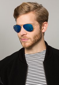Occhiali da sole aviator con lenti riflettenti blu, montatura in metallo dorato e aste sottili, indossati da una persona con una giacca bomber nera e una maglietta a righe.