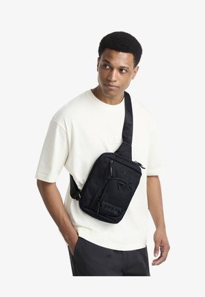 Zwarte crossbodytas met meerdere ritsvakken. Gemaakt van duurzaam stof, met een logo en verstelbare band. Draagt over een licht overhemd.