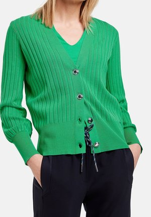Femme portant un cardigan côtelé vert avec des boutons argentés sur un haut vert assorti et un pantalon noir, les mains dans les poches.