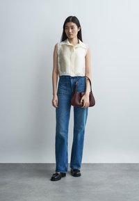 Νέα γυναίκα στέκεται κρατώντας μια σκουρόκαφέ τσάντα, φορώντας μια αμάνικη κρεμ μπλούζα με δαντέλα, μπλε τζιν και μαύρα μοκασίνια.