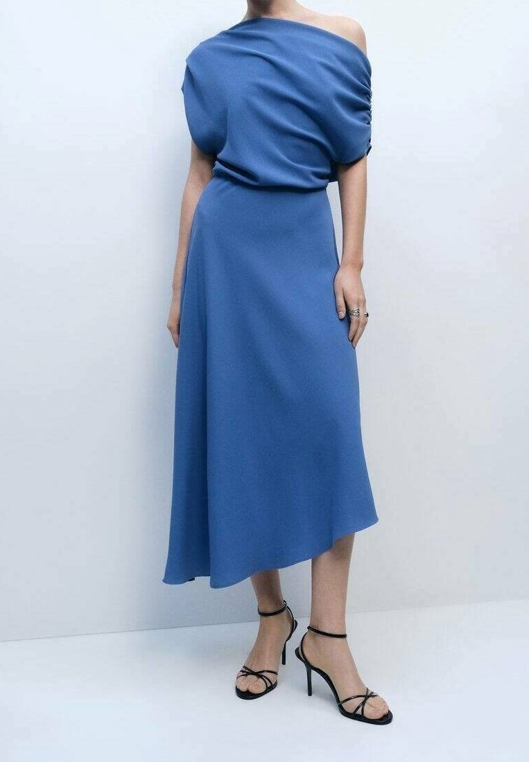 Femme portant une robe midi bleue asymétrique à une épaule avec des sandales à talons hauts noires à brides, debout contre un mur uni.
