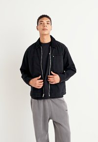 Svart denimjakke med glidelås og krage, med en brystlomme og subtile sømdetaljer, båret over en svart t-skjorte og grå bukser.