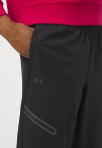 Käsi mustade Under Armour spordipükste taskus, millel on lukuga detail, koos erksa roosa ülaosaga.
