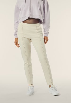 Pantaloni elasticizzati crema con due cuciture verticali, abbinati a un hoodie cropped lilla. Il modello indossa sneakers sportive bianche.