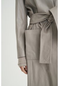 Jasnoszara skórzana kurtka z paskiem w talii, z boczną kieszenią i gładką fakturą. Długie rękawy z dzianinowym paskiem przy mankietach.