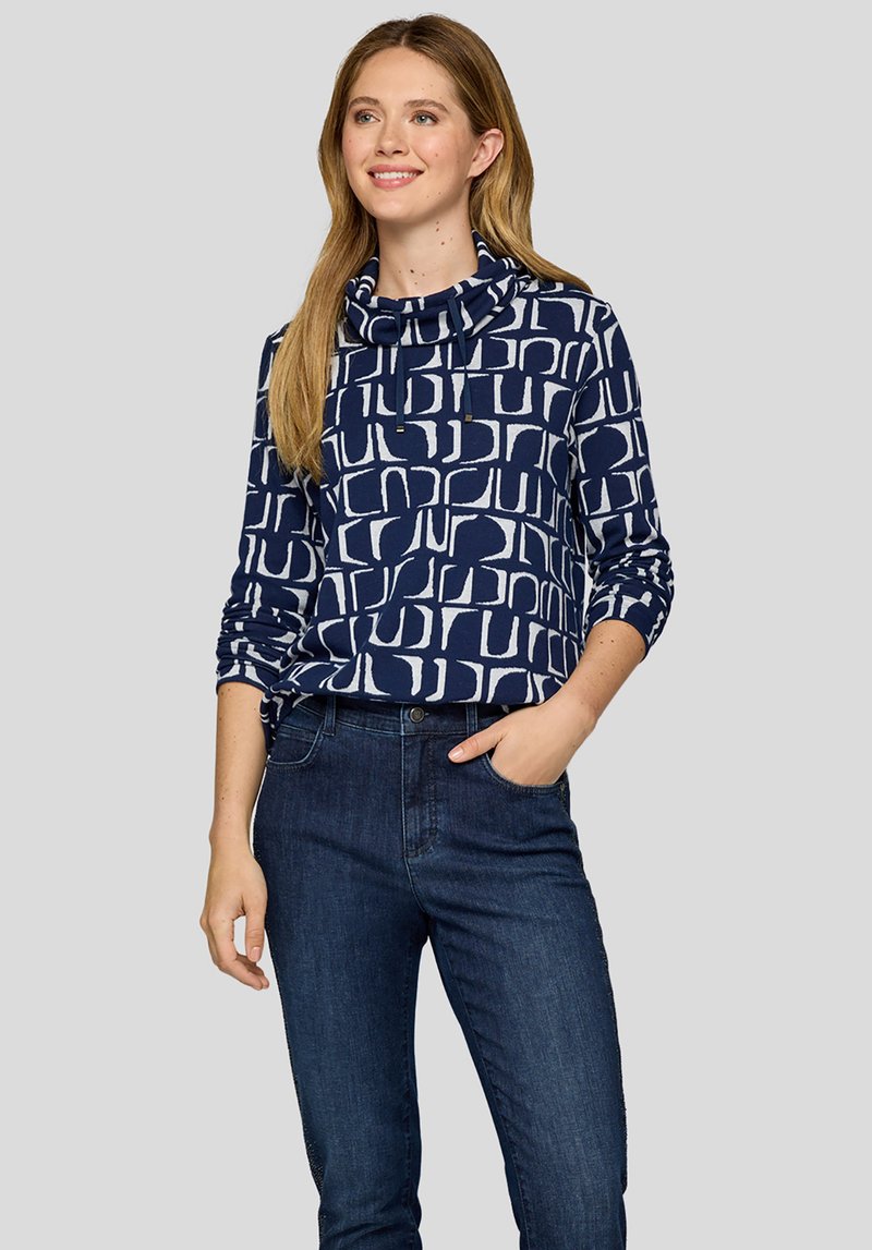 Junge Frau mit langen blonden Haaren, die einen marineblauen Pullover mit Muster und Kordelzugkragen sowie dunkelblaue Jeans trägt und lächelt.