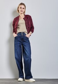 Bordeauxfarbene Bomberjacke, beigefarbenes Strickoberteil, hochgeschnittener, dunkelblauer weiter Jeans, weiße Sneakers, vor einem hellgrauen Hintergrund stehend.