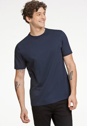 Marineblauw T-shirt met korte mouwen van zachte katoen met een ronde hals. Beschikt over een relaxte pasvorm en een gladde textuur. Zwarte denim jeans gedragen.