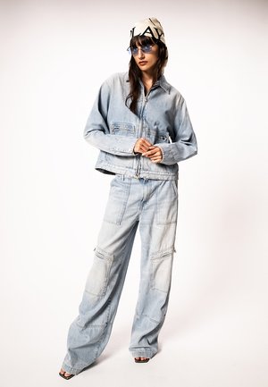 Donna che indossa una giacca di jeans azzurro chiaro e jeans cargo a gamba larga, abbinati a un foulard con motivo e occhiali da sole con lenti sfumate blu.