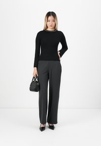 Woman standing, wearing black ribbed sweater, wide-leg black pants, black pointed shoes, holding a small black handbag against white background.