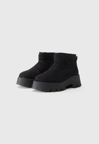 Bottines en daim noir avec des semelles en caoutchouc épaisses et striées, orteils arrondis et cols matelassés sur fond blanc.