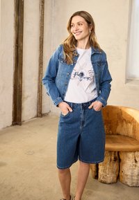 Nő mosolyog, kék farmerdzsekit és térdig érő farmer rövidnadrágot visel, fehér grafikás pólóban, egy helyiségben áll, ahol fa szék és betonpadló található.