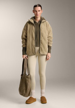 Nő áll, bézs polárdzsekit visel olívazöld kapucnis pulóver fölött, krémszínű leggingset, barna slip-on cipőt, és egy nagy olívazöld táskát tart a kezében.