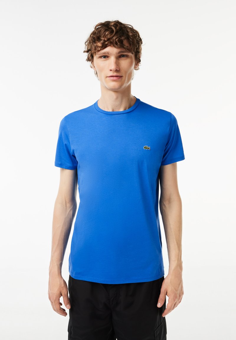 Ung mand med krøllet hår iført en lys blå Lacoste t-shirt og sorte shorts, stående mod en ensfarvet hvid baggrund.
