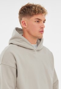 Hanorac gri deschis dintr-un material moale, cu o glugă spațioasă, umeri lăsați și cusături minime pentru o potrivire lejeră.