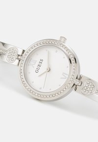 Silver klocka med runt urtavla, vit urtavla, romerska siffror och en kristallbeströdd bezel. Armbandet har hjärtformade detaljer.