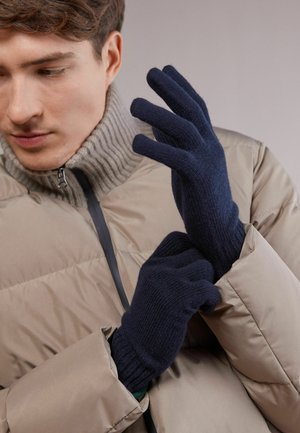 Marineblå strikkede vanter med ribkant, båret over en beige pufferjakke. Blød tekstur, tætsiddende design fremhæver hænderne i en neutral indstilling.