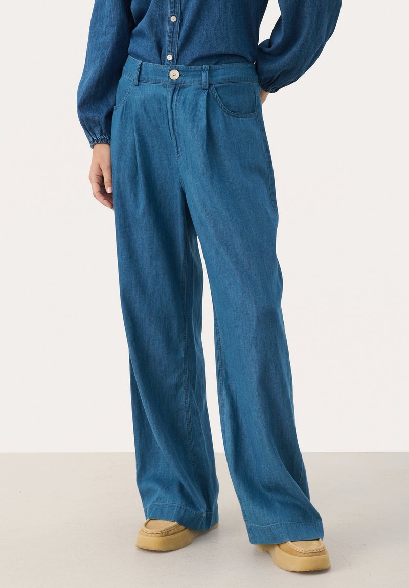 Personne portant un pantalon en jean bleu ample avec un bouton et une fermeture éclair, assorti à des chaussures style mocassin beige et une chemise bleue assortie.