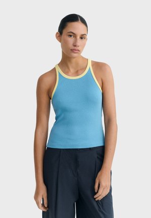 Woman with dark hair in a blue tank top with yellow trim and loose black pants standing against a plain light background.