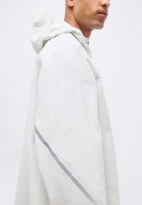 Άτομο που φοράει λευκό μπουφάν με κουκούλα, με αντανακλαστική διαγώνια ρίγα και ανάγλυφο λογότυπο "TERREX" στο πάνω μέρος του μανικιού.