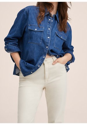 Camisa de denim en azul oscuro con dos bolsillos en el pecho, botonadura frontal y mangas enrolladas, combinada con pantalones ajustados de color crema claro.