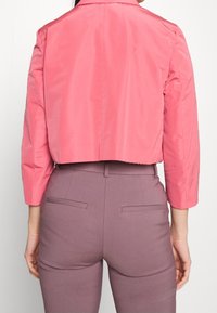 Korte jas in levendig roze, met schoudernaden, lange mouwen en een gladde textuur. Draagt over aangesloten paarse broek.