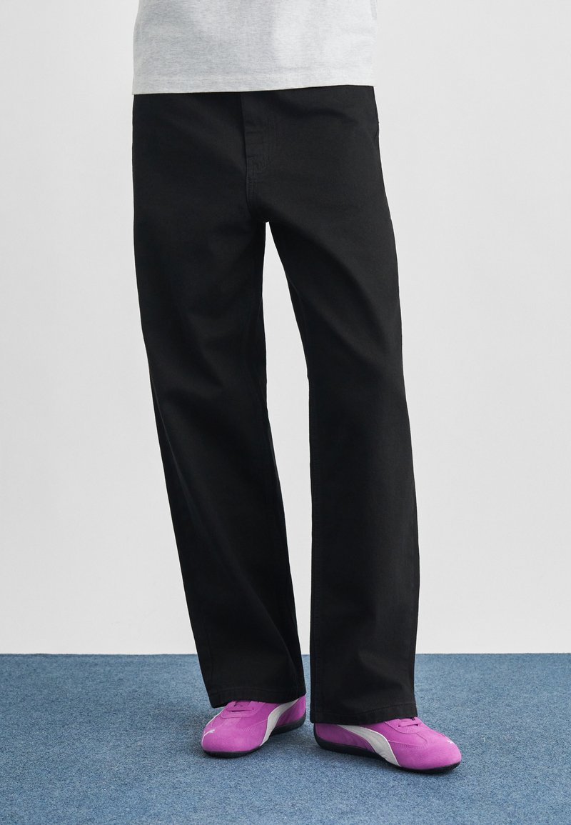 Μαύρο βαμβακερό παντελόνι με άνετη γραμμή, ίσια μπατζάκια και απαλή υφή, συνδυασμένο με παπούτσια ανοιχτού μωβ χρώματος με λευκές λεπτομέρειες.