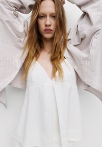 Witte mouwloze top met een V-hals en geplooide stof, gelaagd onder een licht gestreepte oversized blouse met knopen. Gestructureerde, casual uitstraling.