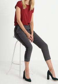 Blusa corta de manga corta roja combinada con pantalones ceñidos de patrón a cuadros gris, con rayas laterales negras, complementada con zapatos de tacón altos verdes.