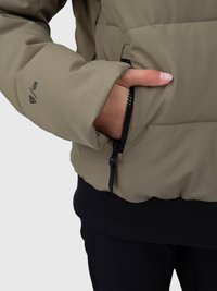 Pessoa a usar uma jaqueta bege acolchoada com um bolso com fecho, mão dentro do bolso, e calças pretas visíveis.