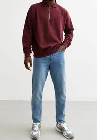Polo a maniche lunghe bordeaux con zip a un quarto, abbinato a jeans in denim azzurro chiaro e sneakers grigie con dettagli bianchi e lacci.